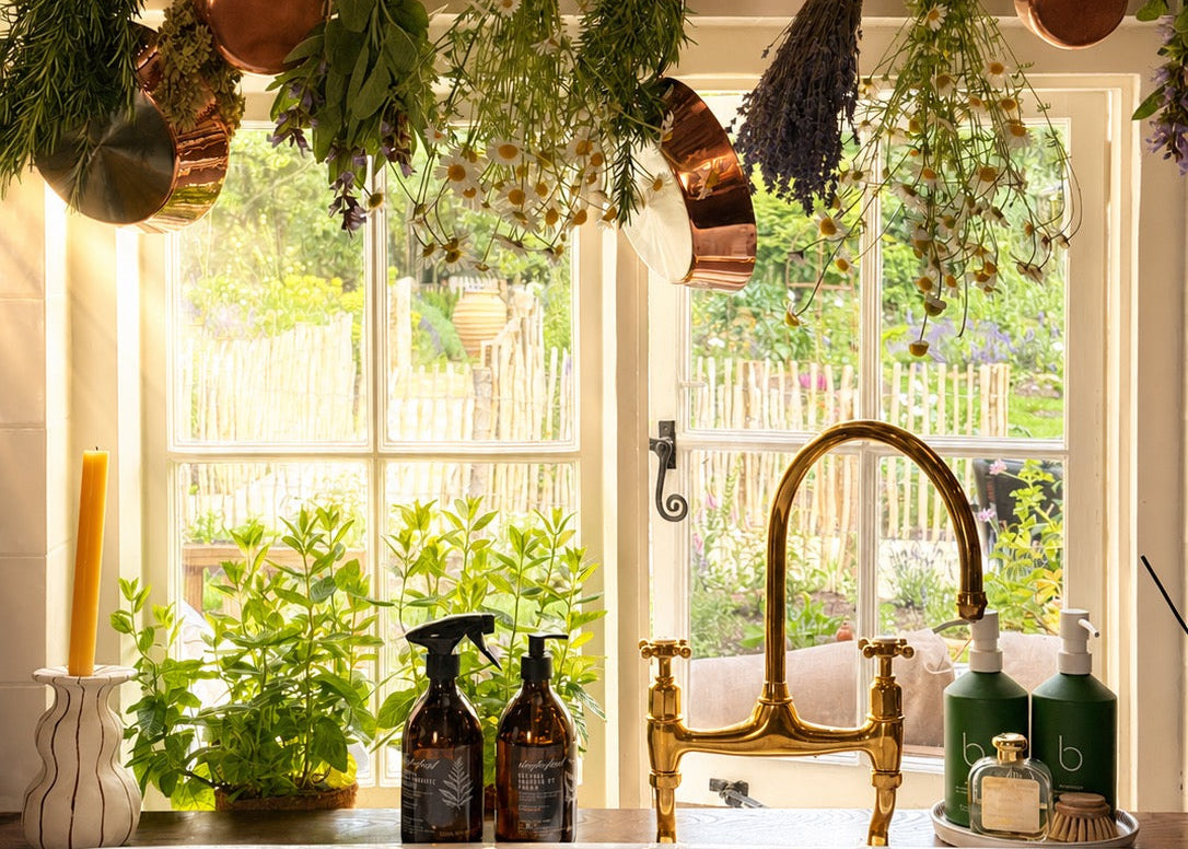 A rustic kitchen scene with a white double-bowl farmhouse sink, brass faucet, hanging copper pots, herbs, and a window looking out onto greenery.
