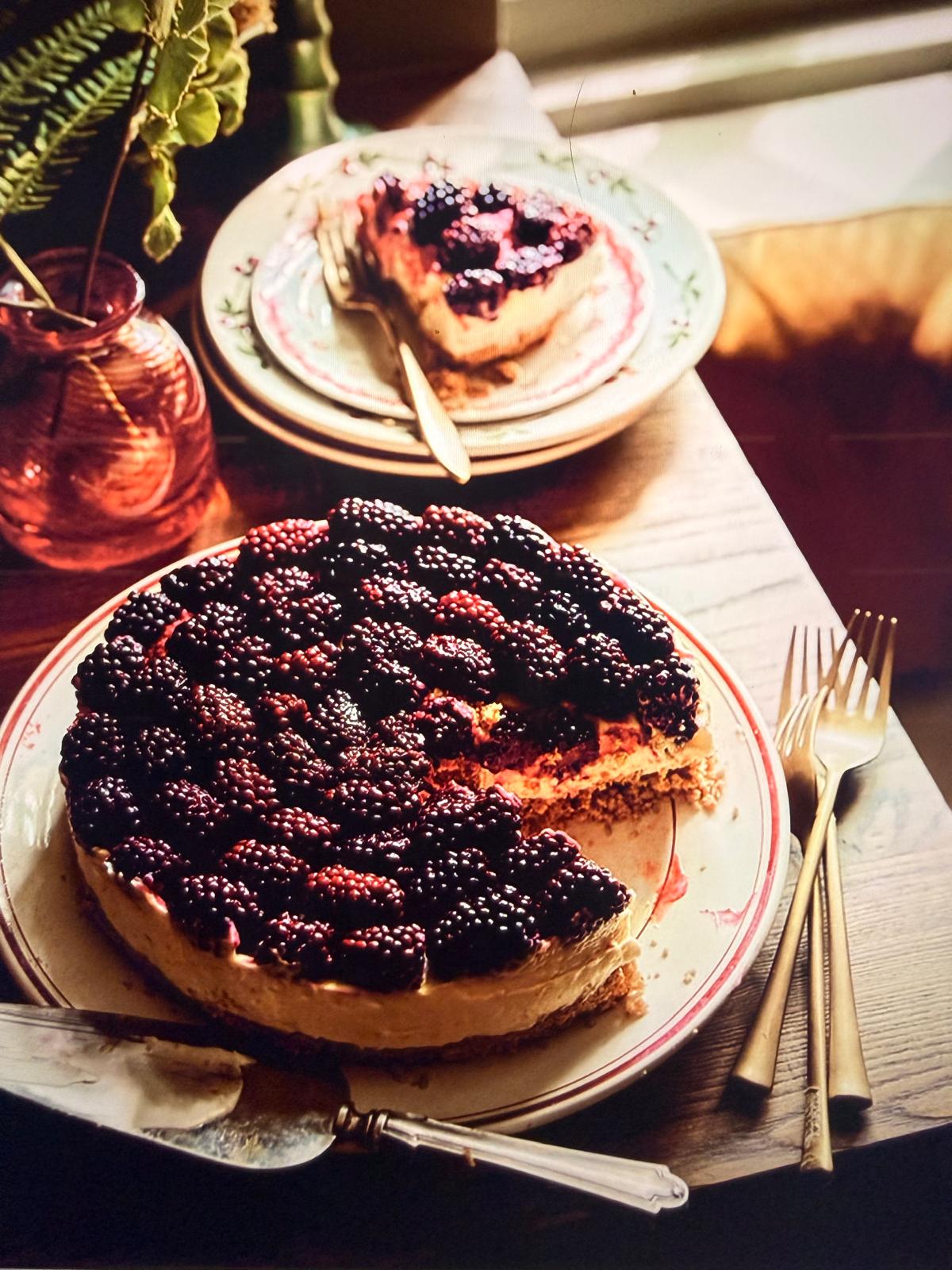 A large round berry cake topped with fresh blackberries sits on a wooden table, with a slice cut and placed on a plate in the background. Gold-toned forks, a pink glass vase with greenery, and warm natural light create an inviting dining atmosphere.