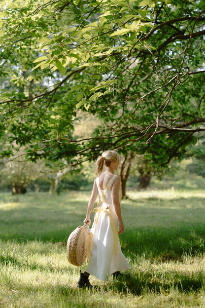 Woman in a white dress holding a straw bag in a park with trees and grass.