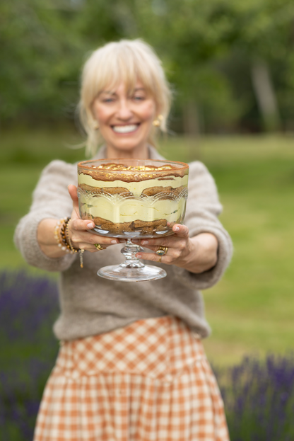 Clodagh McKenna in a beige sweater and peach-and-white gingham skirt holds a glass trifle with her pistachio tiramisu dessert platter outdoors in a garden setting with blurred greenery and purple flowers in the background.
